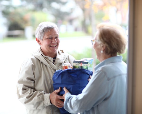 Eine ältere Frau reicht einer anderen älteren Frau eine Tasche mit Lebensmitteln | © Caritas München und Oberbayern